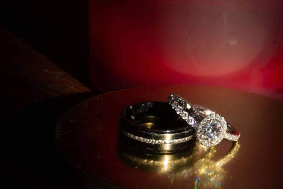 Two rings, one gold band and one silver with a large diamond, resting on a reflective surface.